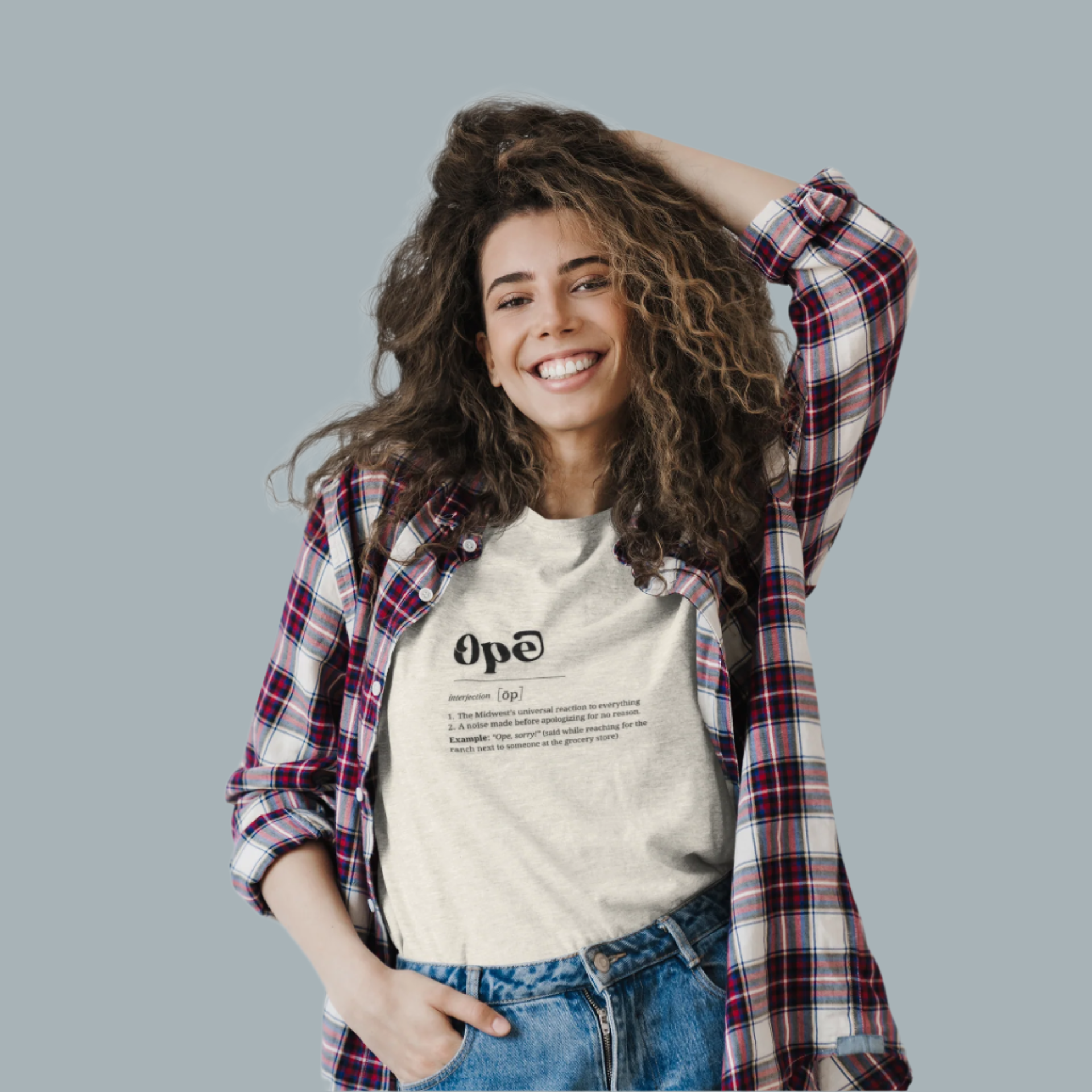 A woman smiling and wearing a beige crew neck t-shirt with the word 'Ope' printed on it, paired with a plaid shirt and jeans.