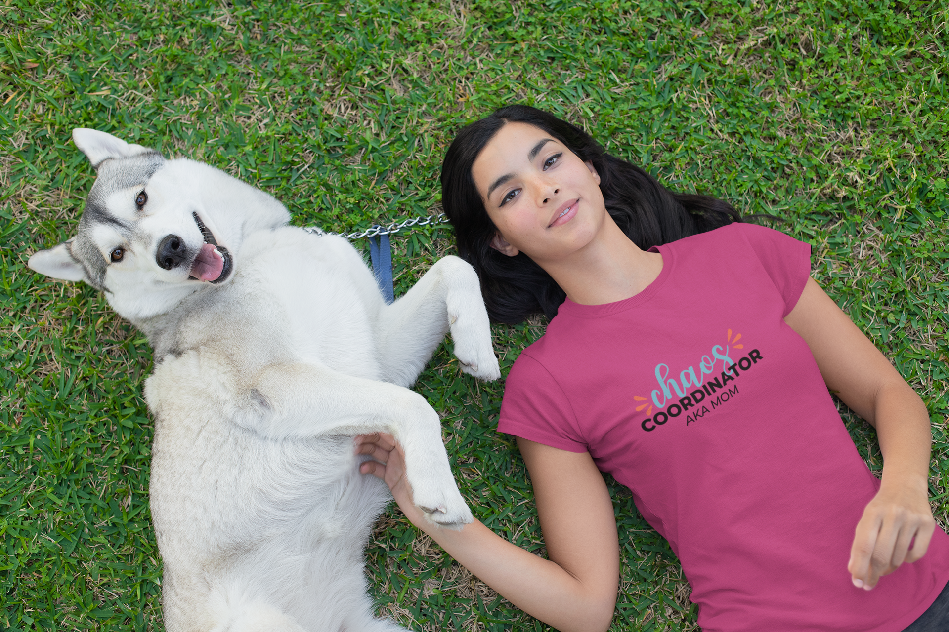 Woman lying on grass with a white dog, wearing a pink t-shirt with text.